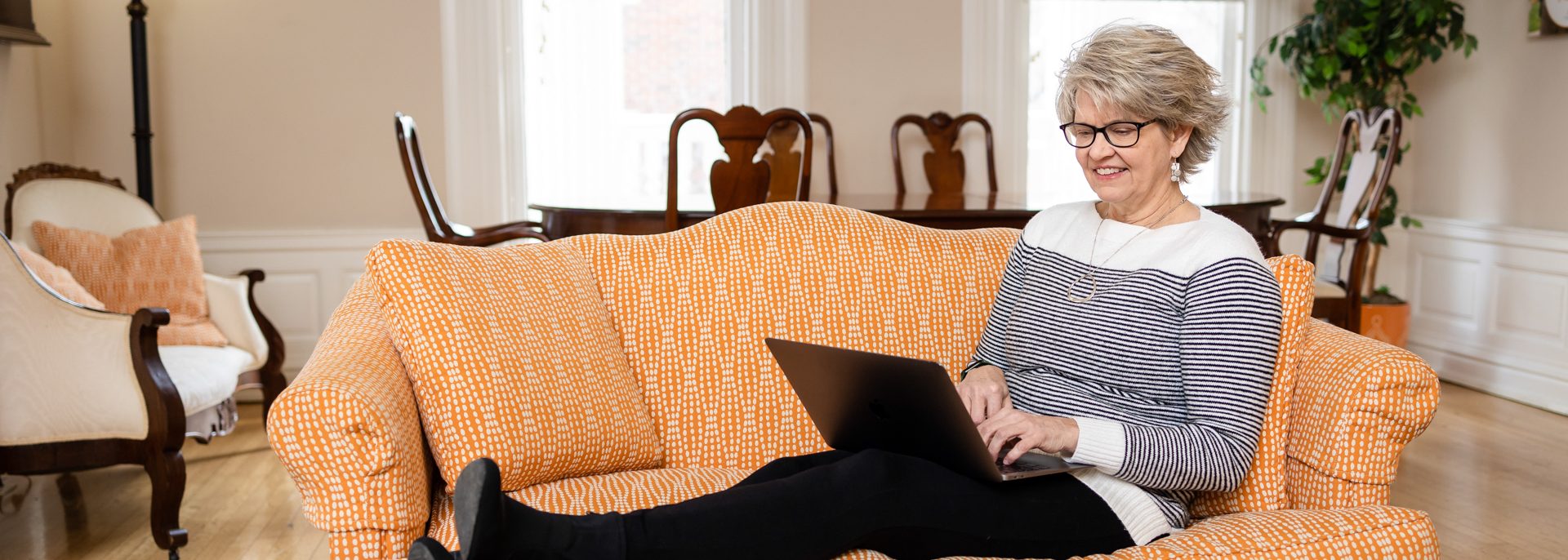 older female CSW online student sitting on orange couch and working on laptop