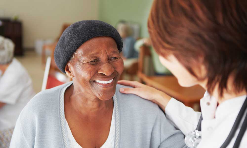 A care provider in the foreground rests her hand on the shoulder of an smiling elderly woman in center frame.