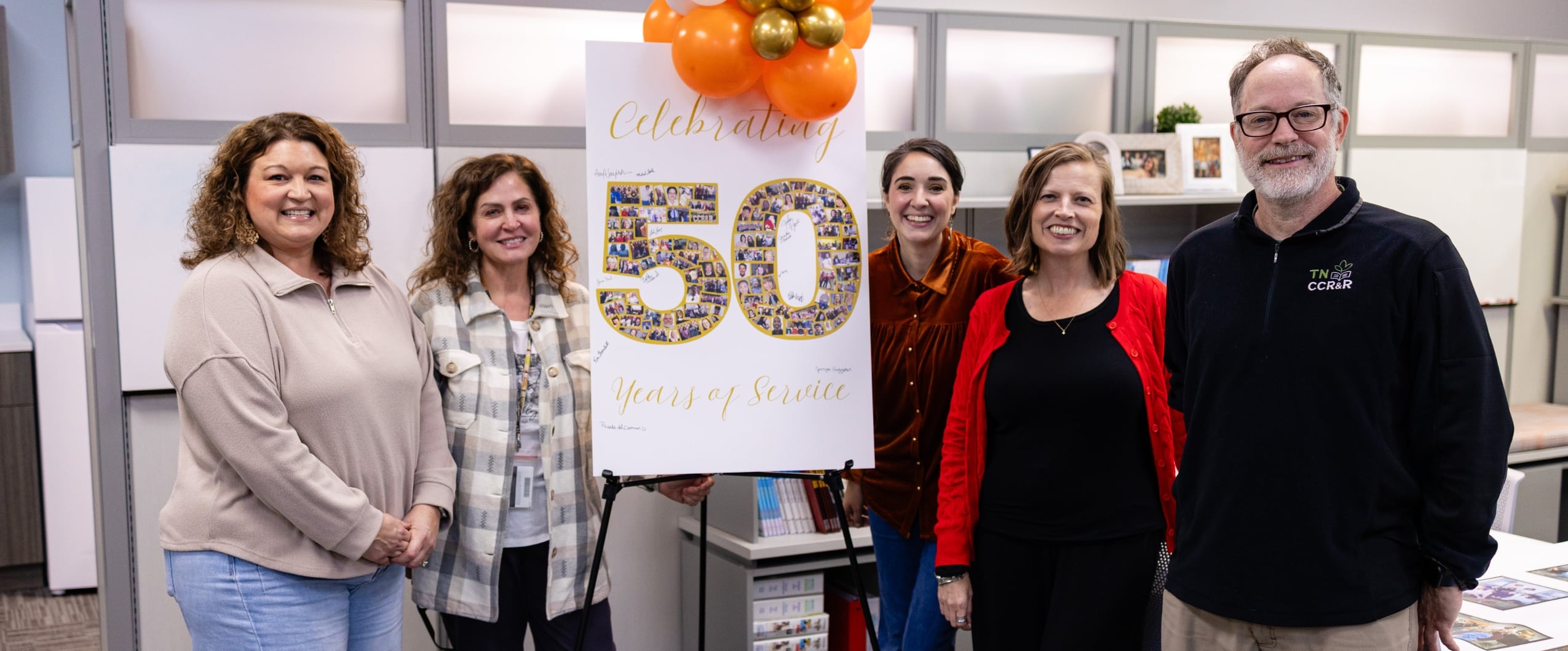 Five smiling colleagues, three women and two men, stand around a large white sign that reads 'Celebrating 50 Years of Service'. The number '50' is a photo collage, and orange and gold balloons are at the top. The man on the right wears a black pullover with a 'TN CCR&R' logo.
