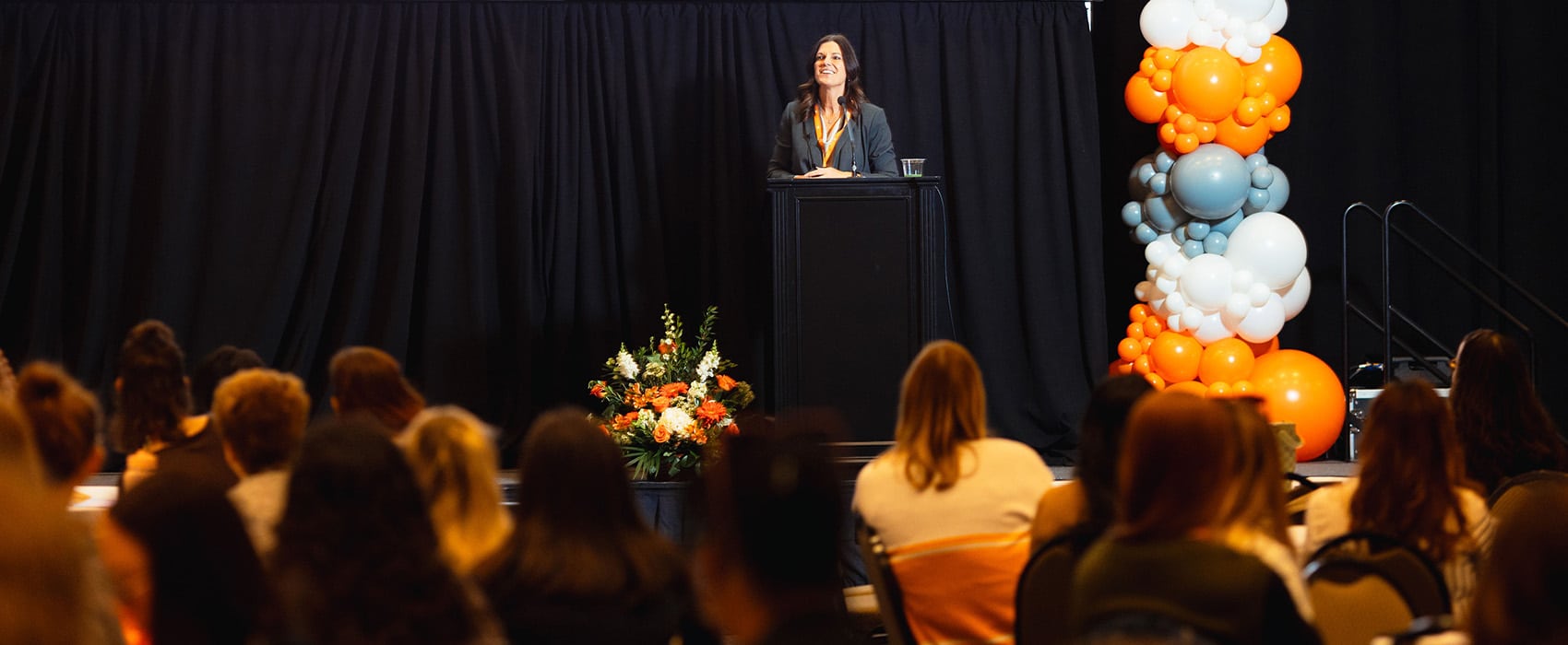 A smiling woman speaks at a podium on a stage, addressing an audience. The stage is decorated with flowers and orange, white, and grey balloons.