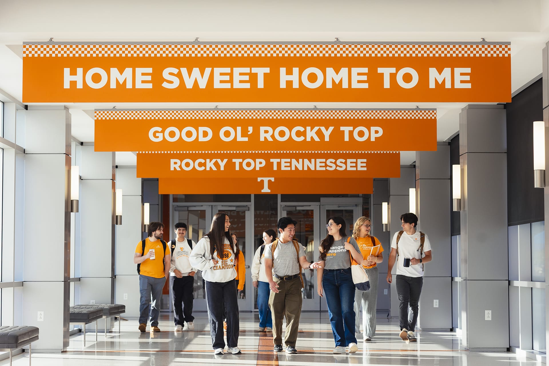 A group of diverse college students walk down a bright, modern hallway. Orange banners overhead read "HOME SWEET HOME TO ME," "GOOD OL' ROCKY TOP," and "ROCKY TOP TENNESSEE," with a 'T' logo.