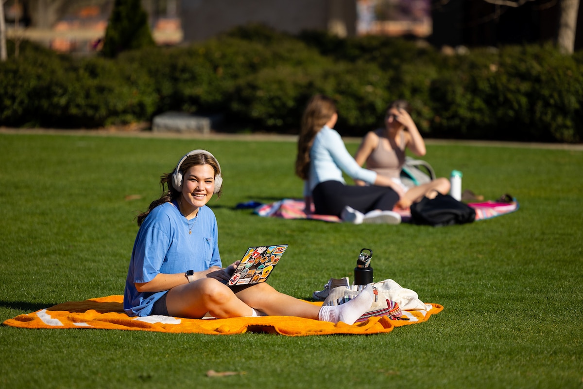 A student smiles for the camera while working on a laptop and laying on a UT blanket at the Humanities Plaza