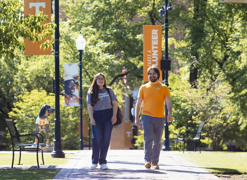 A woman and a man walk on a paved path on a sunny university campus, flanked by trees and banners with the University of Tennessee logo.