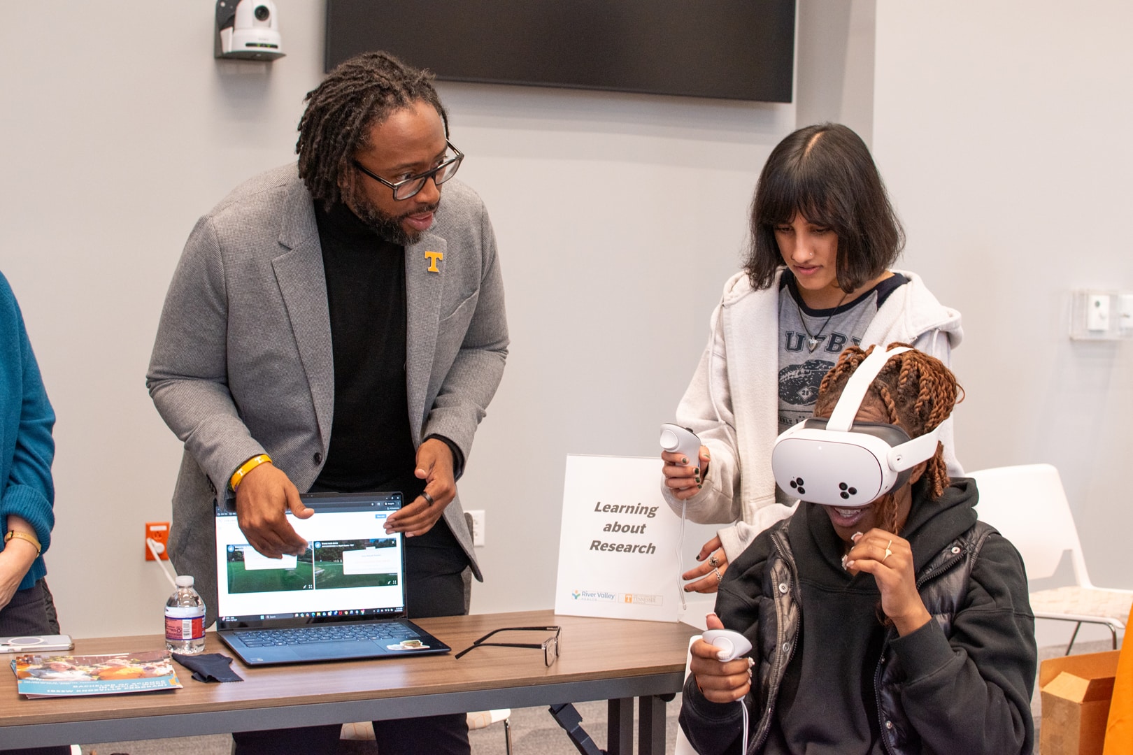 Three people interact with virtual reality and a laptop. One person wears a VR headset, another holds controllers, and a man points at a laptop screen.