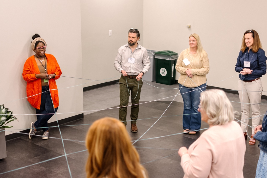 Six people standing in a circle, holding and connecting strands of string that form a web-like pattern on the floor, participating in a therapy training exercise.