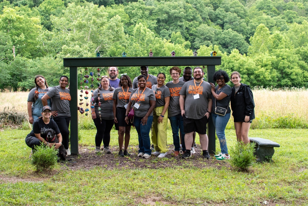 A diverse group of fourteen people and a black dog, mostly wearing gray "SOCIAL WORKER" t-shirts, pose outdoors under a green archway decorated with colorful hanging items and birdhouses.