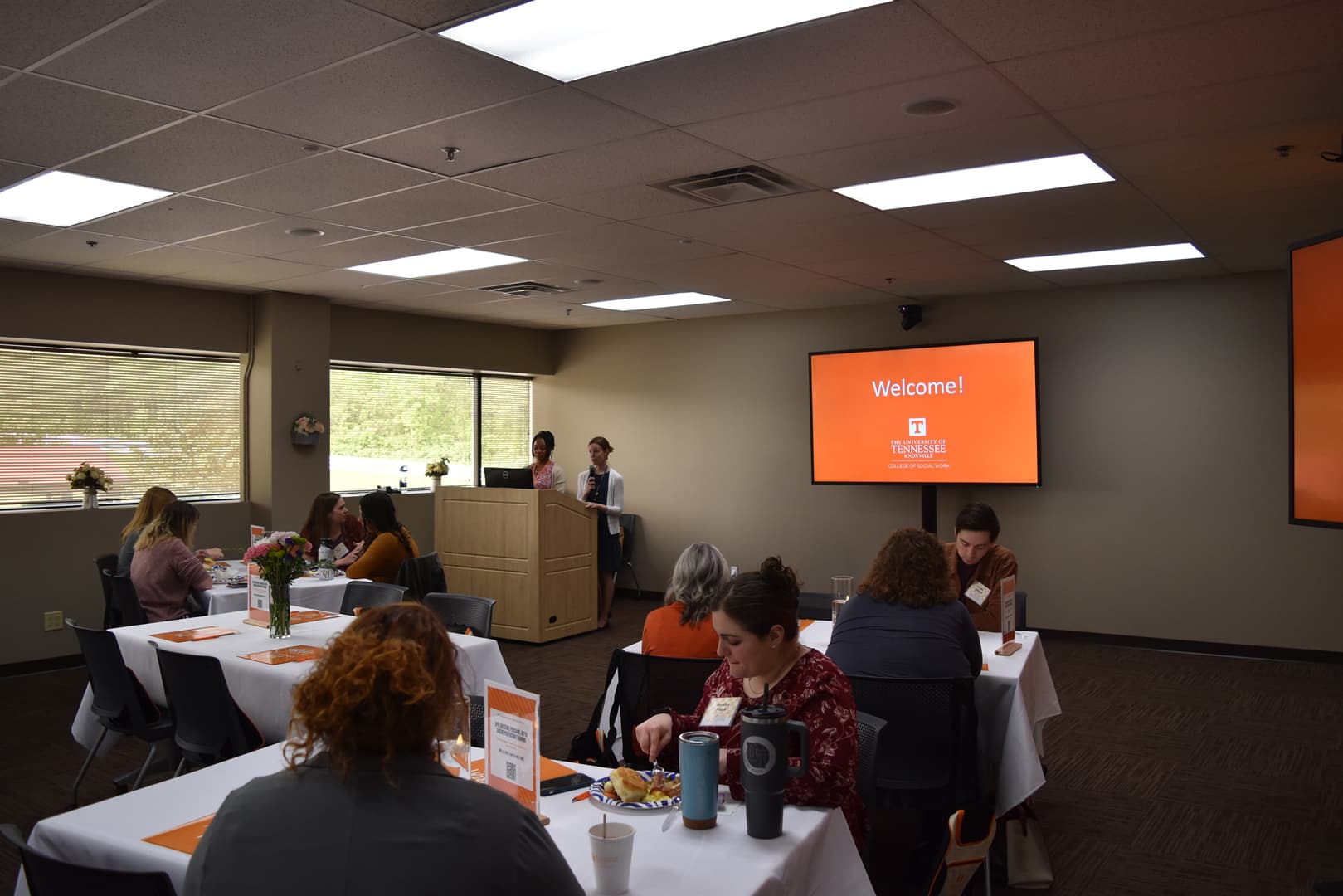 Two women present at a podium to an audience seated at tables in a room with a 'Welcome!' screen from the University of Tennessee, Knoxville College of Social Work.