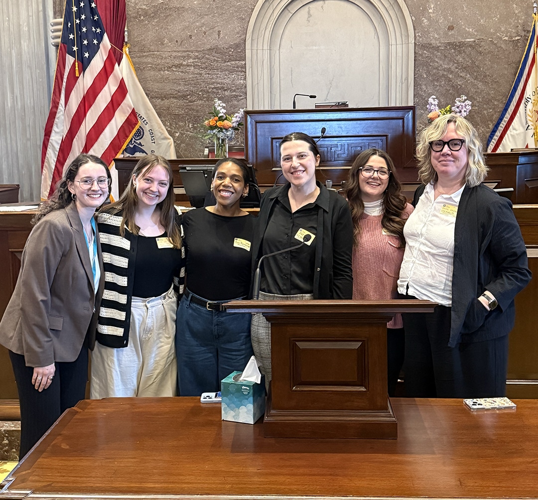 Six smiling women stand behind a wooden podium in a government hall, with American and state flags in the background.