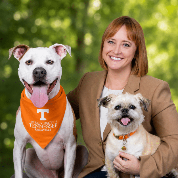 A smiling woman with two dogs. A large white dog wears an orange University of Tennessee bandana, and a small tan dog is held by the woman. All three are looking at the camera with happy expressions.