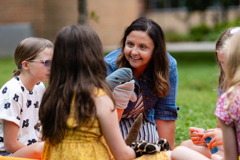 A smiling woman with a shark puppet interacts with several young children sitting on green grass outdoors.