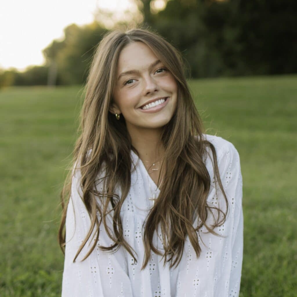 A young woman with long brown hair smiles at the camera, wearing a white eyelet top and gold hoop earrings, against a blurred green and golden background.