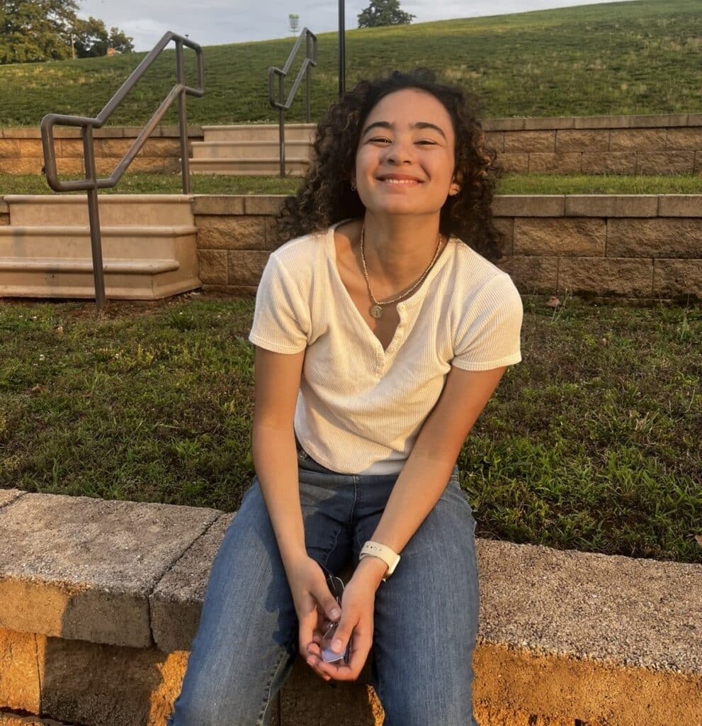 A young woman with curly hair smiles while sitting on a stone wall, with a grassy hill and stone steps in the background.