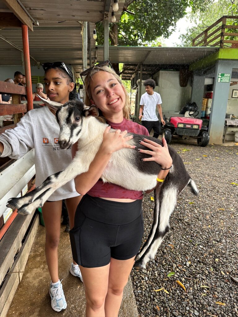Two young women, one holding a black and white baby goat, standing outdoors in the rain.