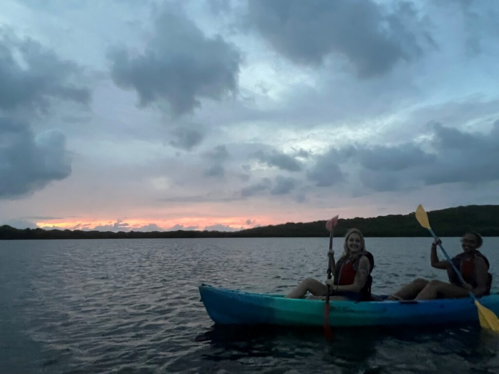 Two people in a blue and teal kayak on a lake at dusk, wearing red life vests and holding paddles, with a colorful sky and dark clouds overhead.