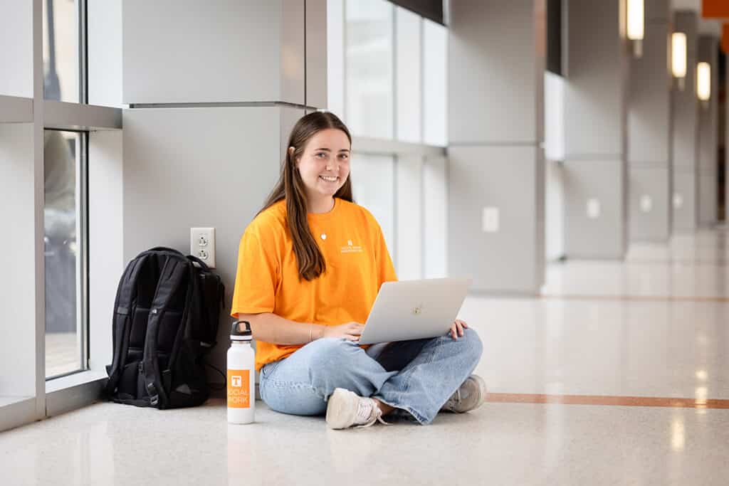 A smiling young woman in an orange 'Social Work Ambassador' t-shirt sits cross-legged on a floor, using a laptop. A backpack and water bottle are beside her in a bright hallway.