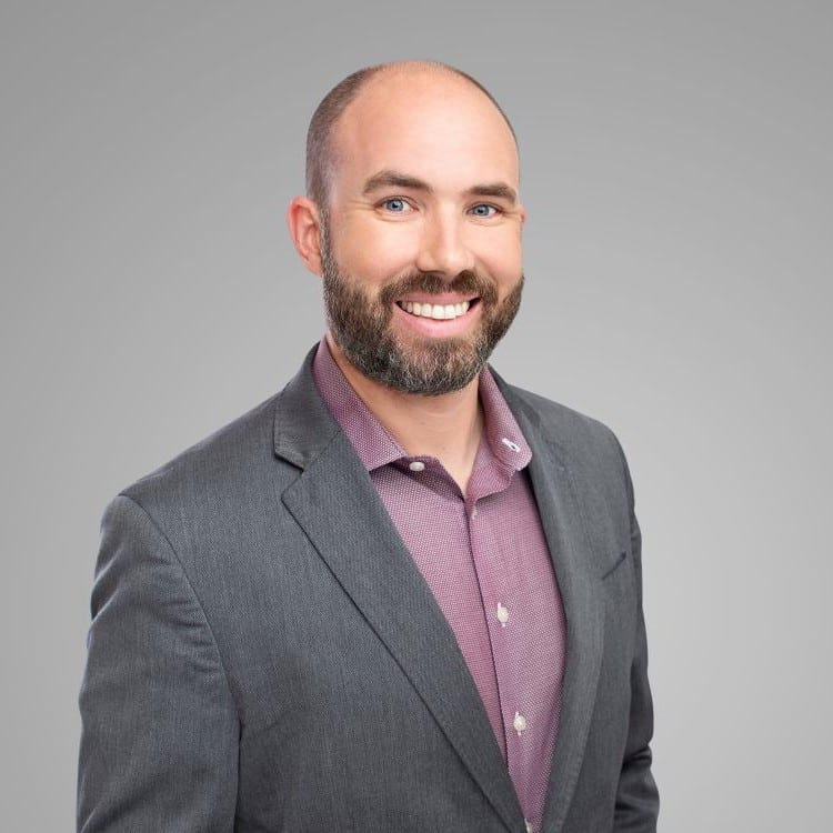 Headshot of a smiling man with a bald head and beard, wearing a grey blazer and maroon shirt.