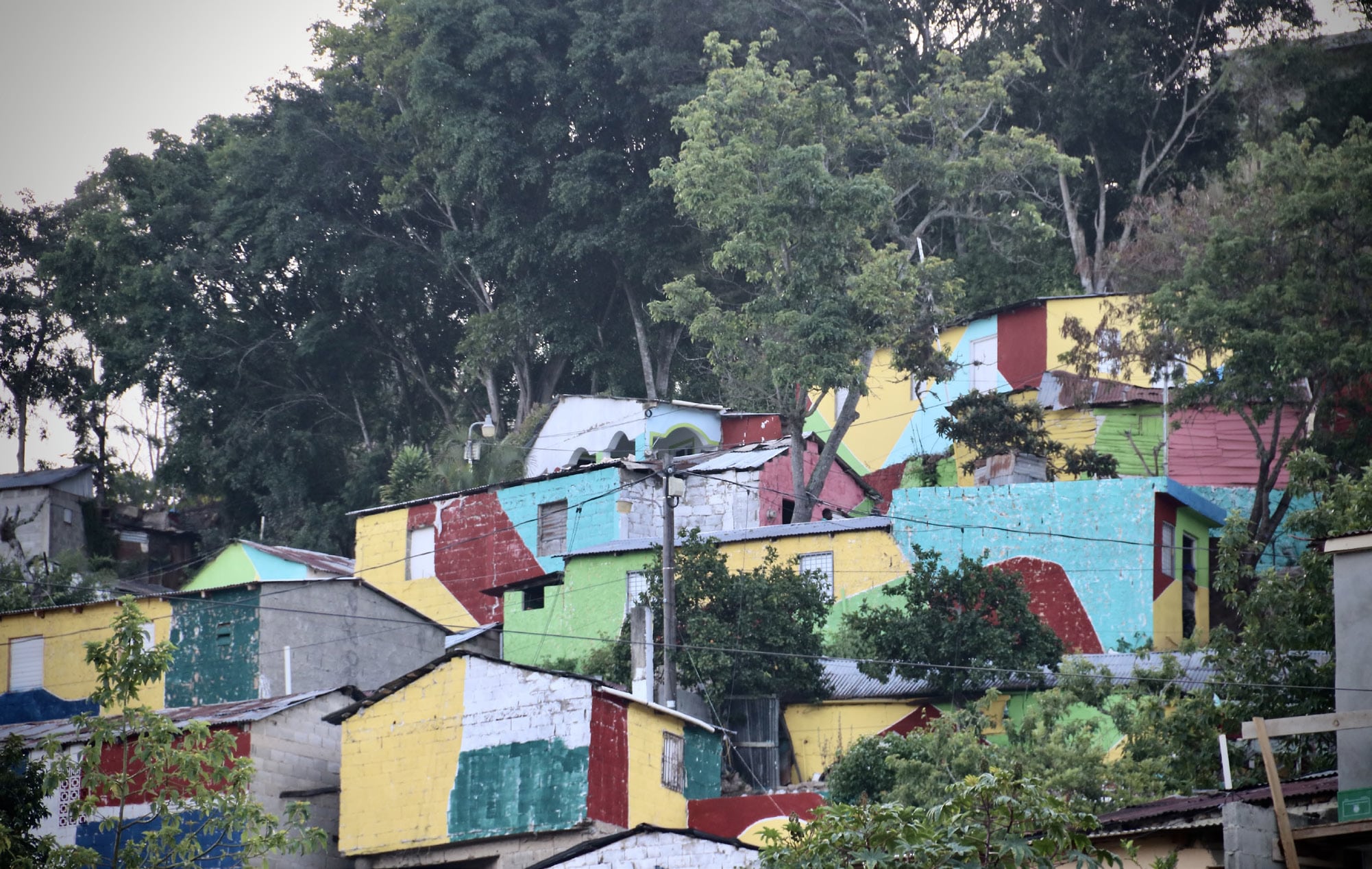 A hillside community with houses painted in bright, geometric blocks of yellow, red, turquoise, green, and white, surrounded by dense green trees.