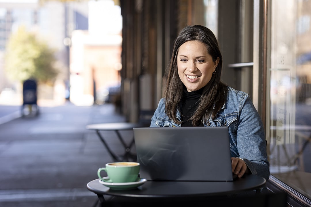 A smiling woman works on a laptop at an outdoor cafe table with a green coffee cup beside her.
