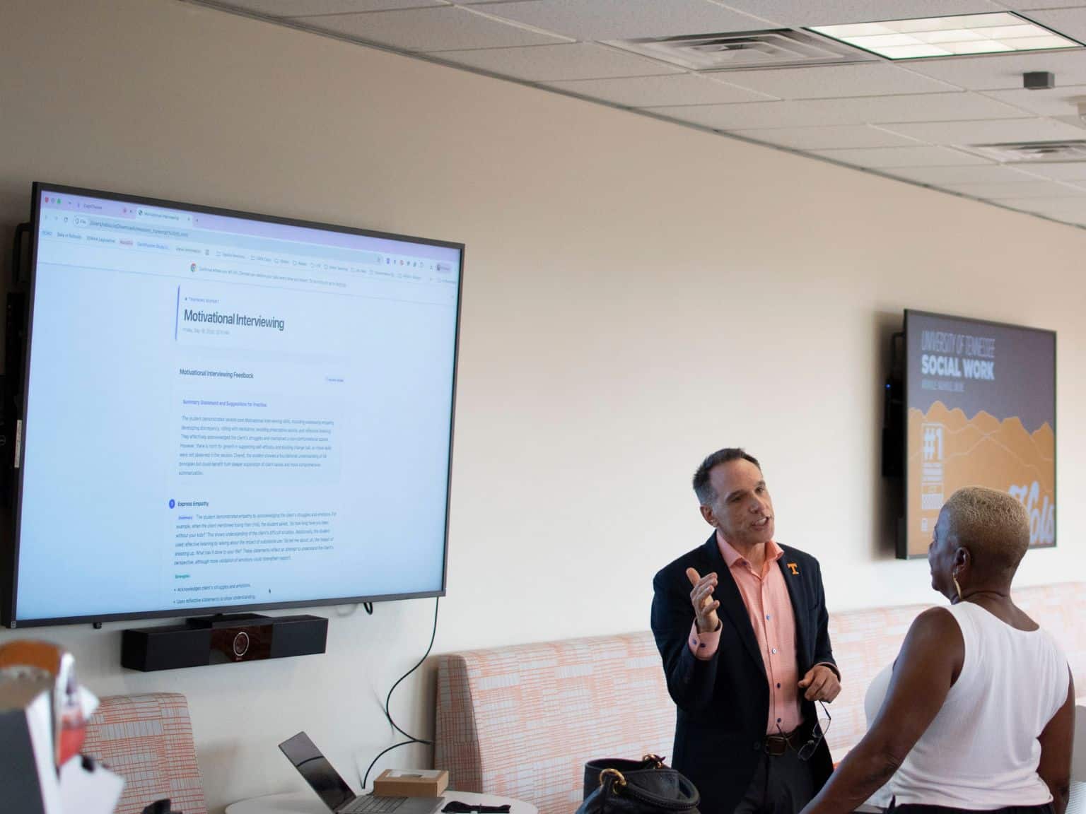 A man and a woman talking in a room with two large screens. One screen displays 'Motivational Interviewing' training material, and the other shows 'University of Tennessee Social Work' branding.