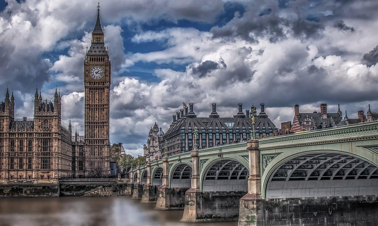 A dramatic view of Big Ben, the Houses of Parliament, and Westminster Bridge over the River Thames under a cloudy sky.