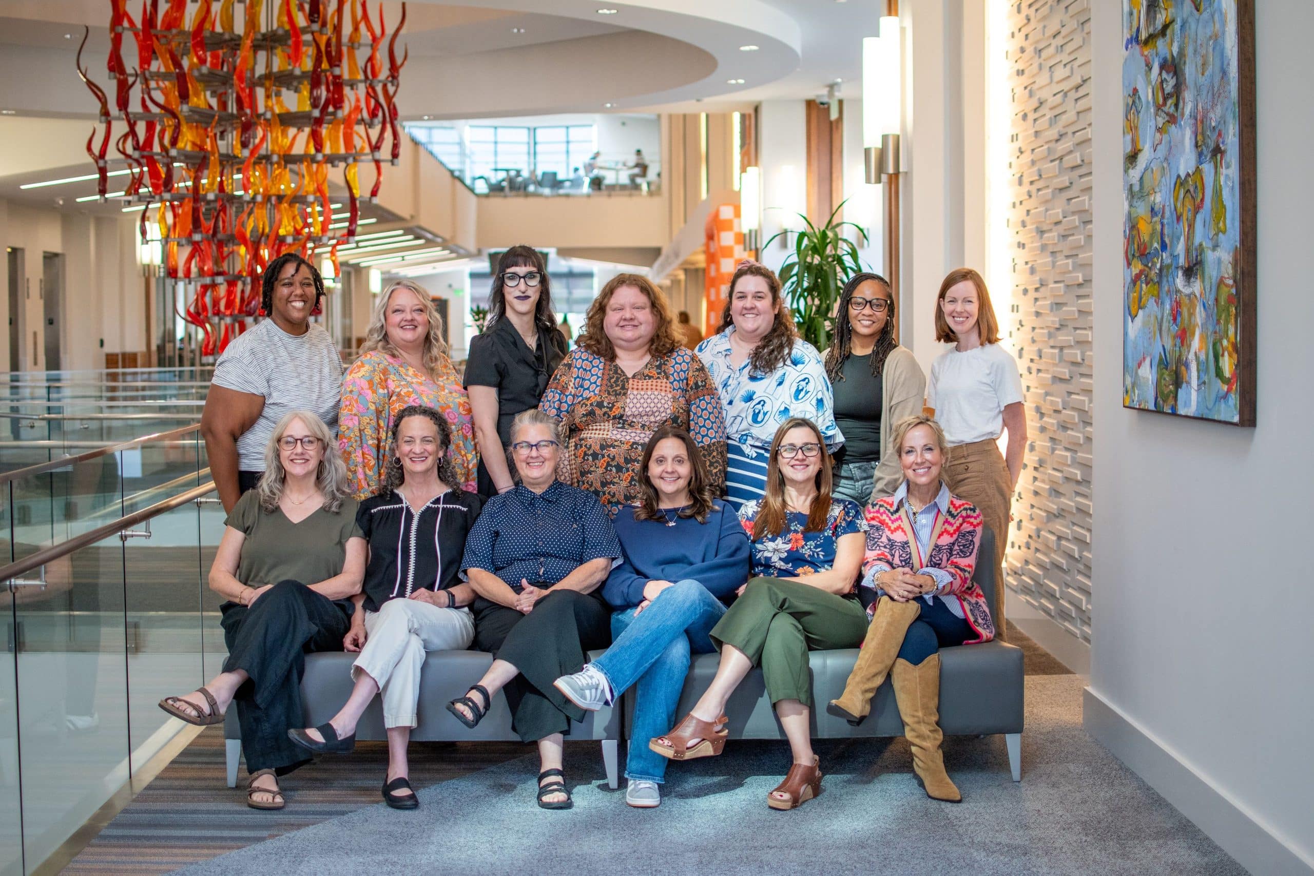 Twelve women, diverse in age and ethnicity, smiling for a group photo in a modern building lobby with a colorful glass sculpture and abstract art.