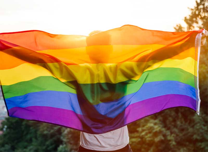 person standing towards sun with rainbow flag behind their back