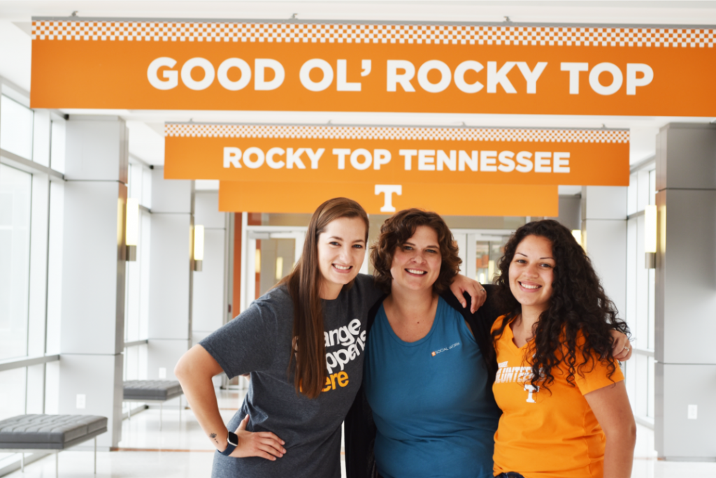 Three woman standing under orange sign with "good ol rocky top" written across it